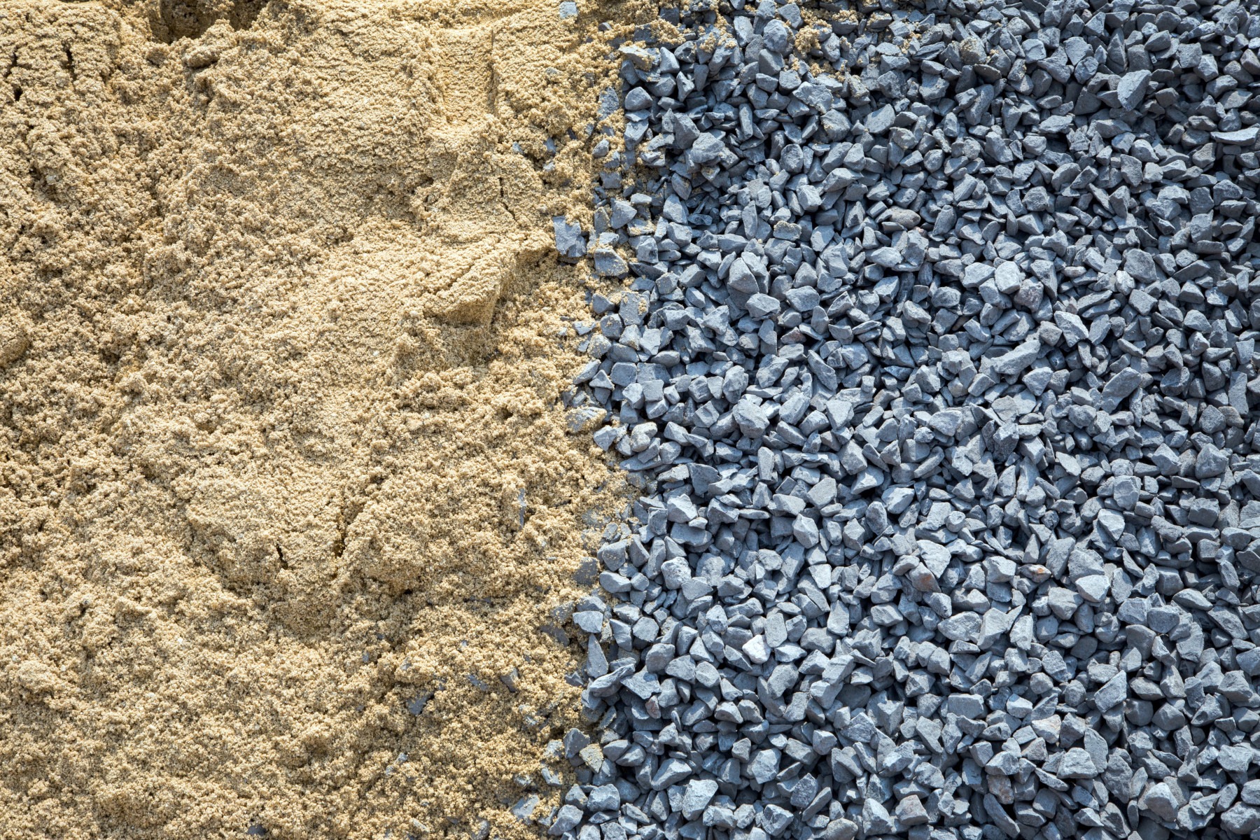Dwie pryzmy materiałów budowlanych: po lewej piasek o drobnej frakcji i jasnym odcieniu beżu, po prawej żwir o ostrych krawędziach i szarym kolorze, wyraźnie oddzielone od siebie