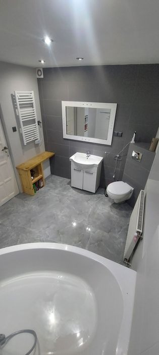 Wykończona łazienka z szarą glazurą, białą armaturą, drewnianą półką i grzejnikiem drabinkowym, widok z perspektywy wanny.