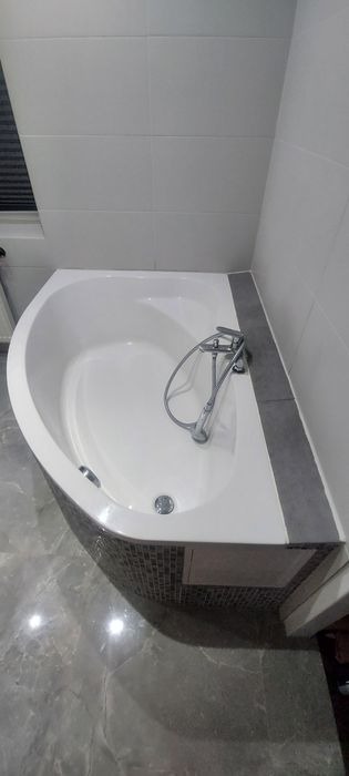 Biała wanna narożna z chromowaną baterią, obłożona mozaiką i szarą płytką, na podłodze szare płytki imitujące marmur, ściany wyłożone białymi płytkami.