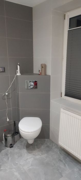 Szara łazienka po remoncie z podwieszaną toaletą, grzejnikiem, roletami w oknie i marmurową podłogą