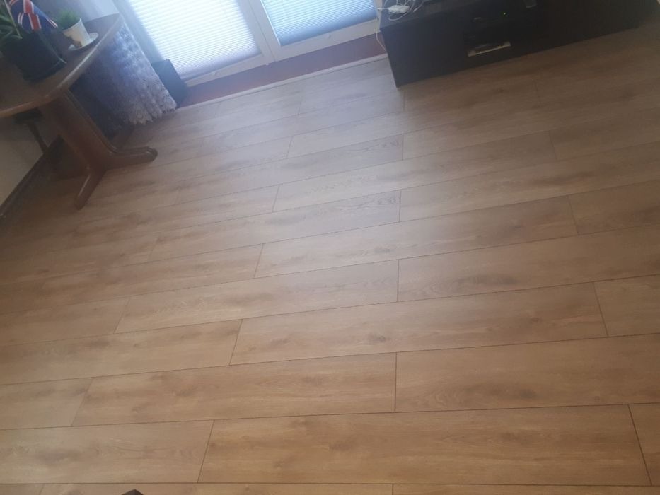 Jasne panele podłogowe ułożone wzdłuż pokoju, widoczny fragment drewnianego stołu z dekoracjami oraz czarna szafka RTV pod oknem z żaluzjami.