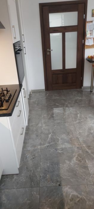 Kuchenna podłoga wyłożona szarymi płytkami imitującymi marmur, widoczny fragment białej zabudowy kuchennej z czarnym blatem i brązowe drzwi z szybami.