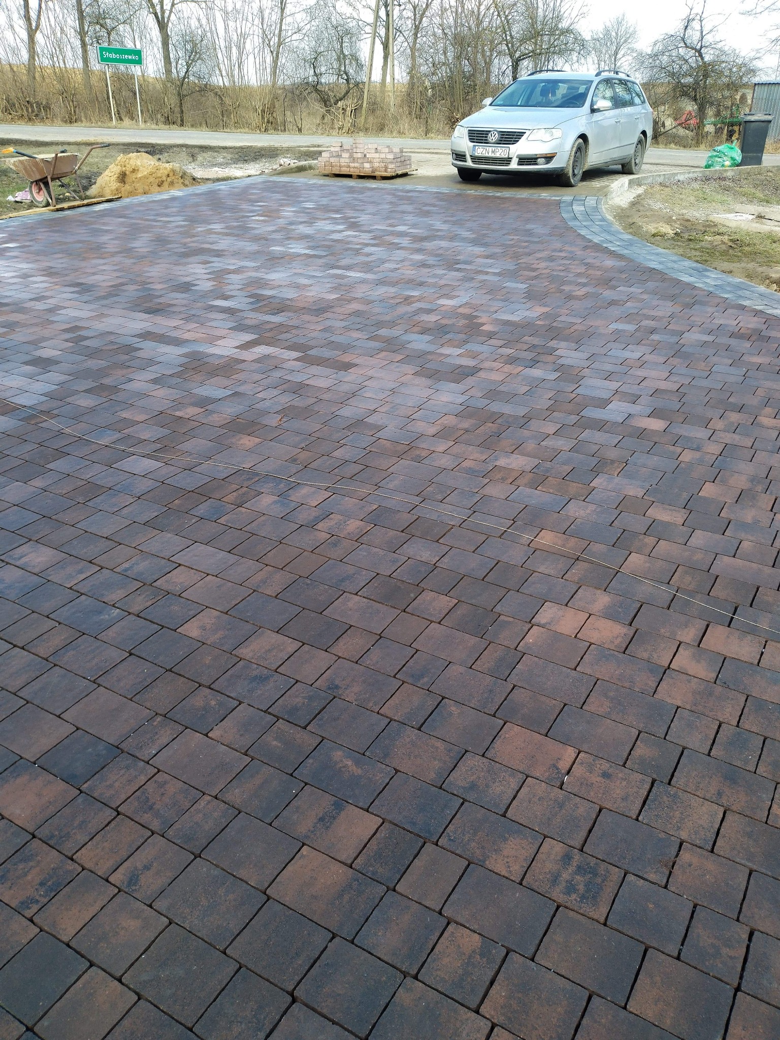 Ułożona kostka brukowa w odcieniach brązu i szarości tworząca podjazd dla samochodu osobowego, widoczny znak miejscowości Słaboszewko i przygotowane materiały budowlane