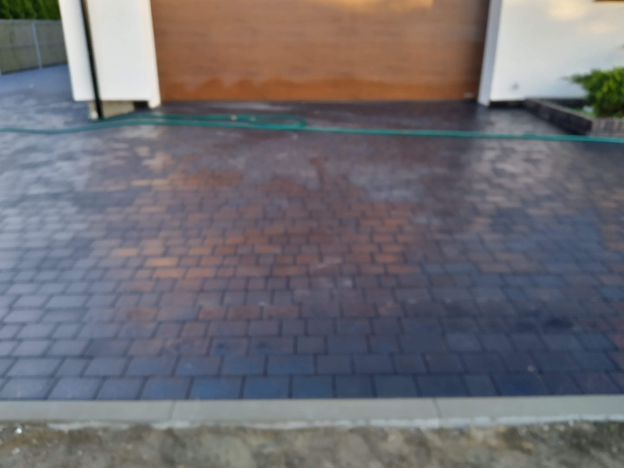 Ułożona kostka brukowa w odcieniach brązu i grafitu przed garażem, widoczny fragment zielonego węża ogrodowego i obrzeże chodnikowe.