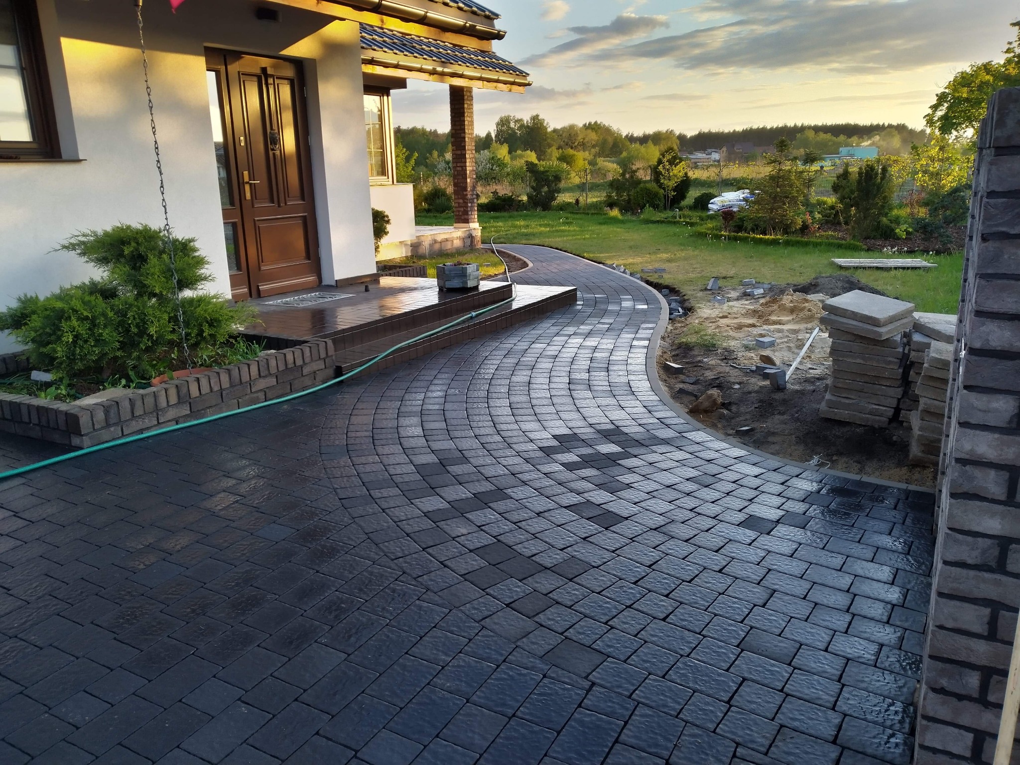 Zakrzywiony chodnik z ciemnoszarej kostki brukowej prowadzący do domu z brązowymi drzwiami, widoczne niedokończone prace i stos kostki obok, w tle zieleń ogrodu i zachodzące słońce.