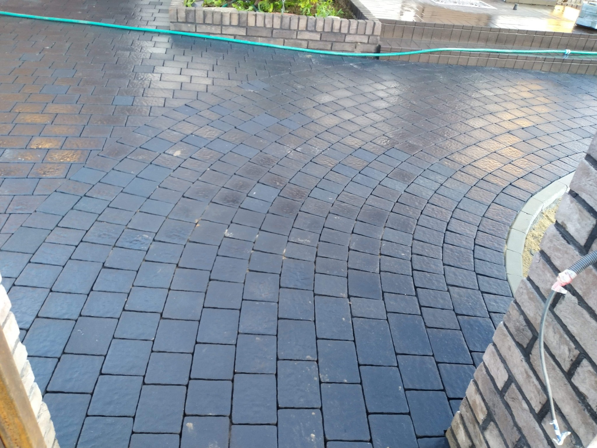 Świeżo ułożona, mokra, grafitowa kostka brukowa ułożona w łukowaty wzór, obok krawężnik i fragment murku z cegły klinkierowej, zielony wąż ogrodowy na górze zdjęcia.