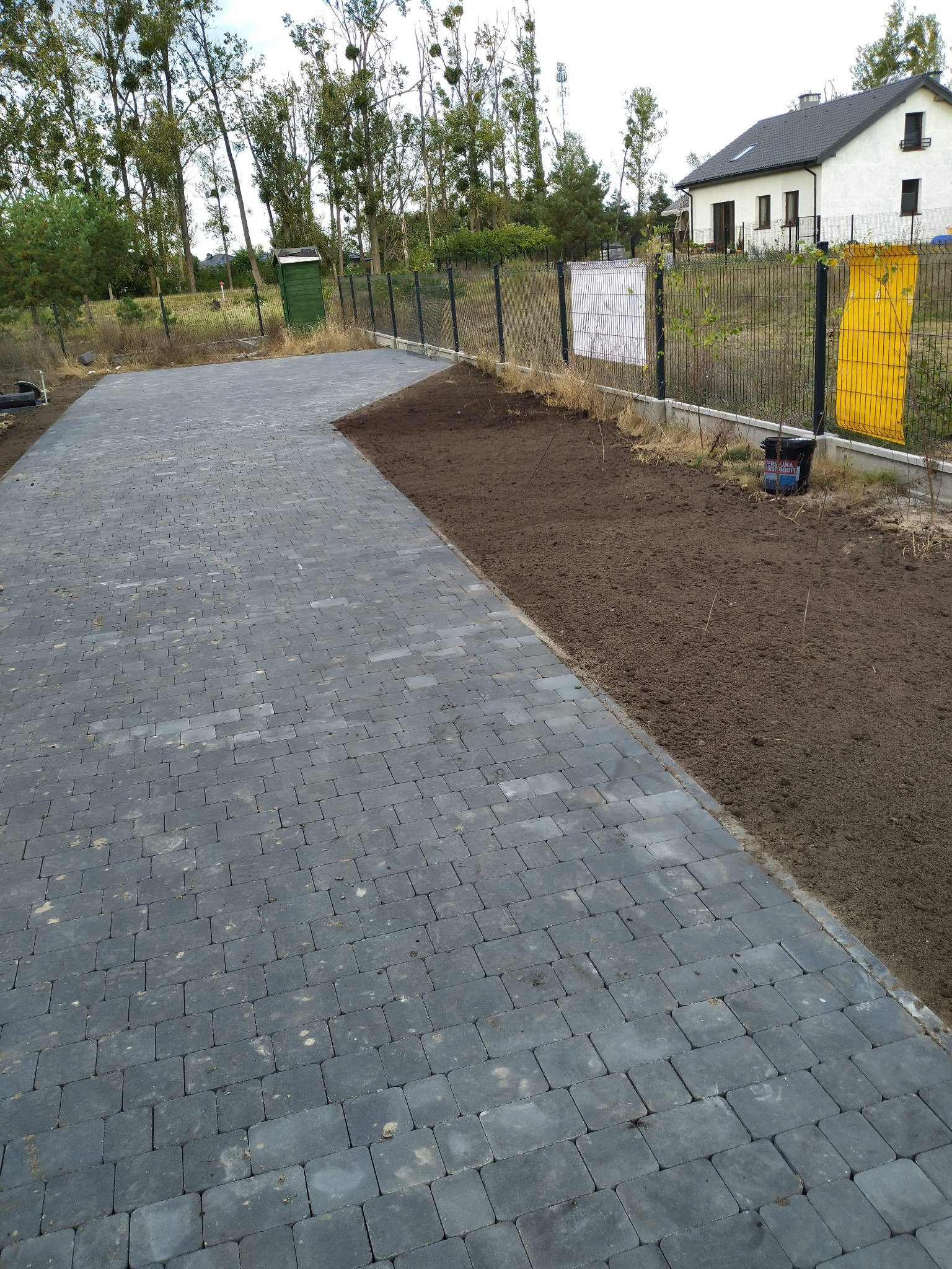 Ułożona szara kostka brukowa na podjeździe, obok świeżo nawieziona ziemia pod trawnik, w tle dom jednorodzinny i drzewa.