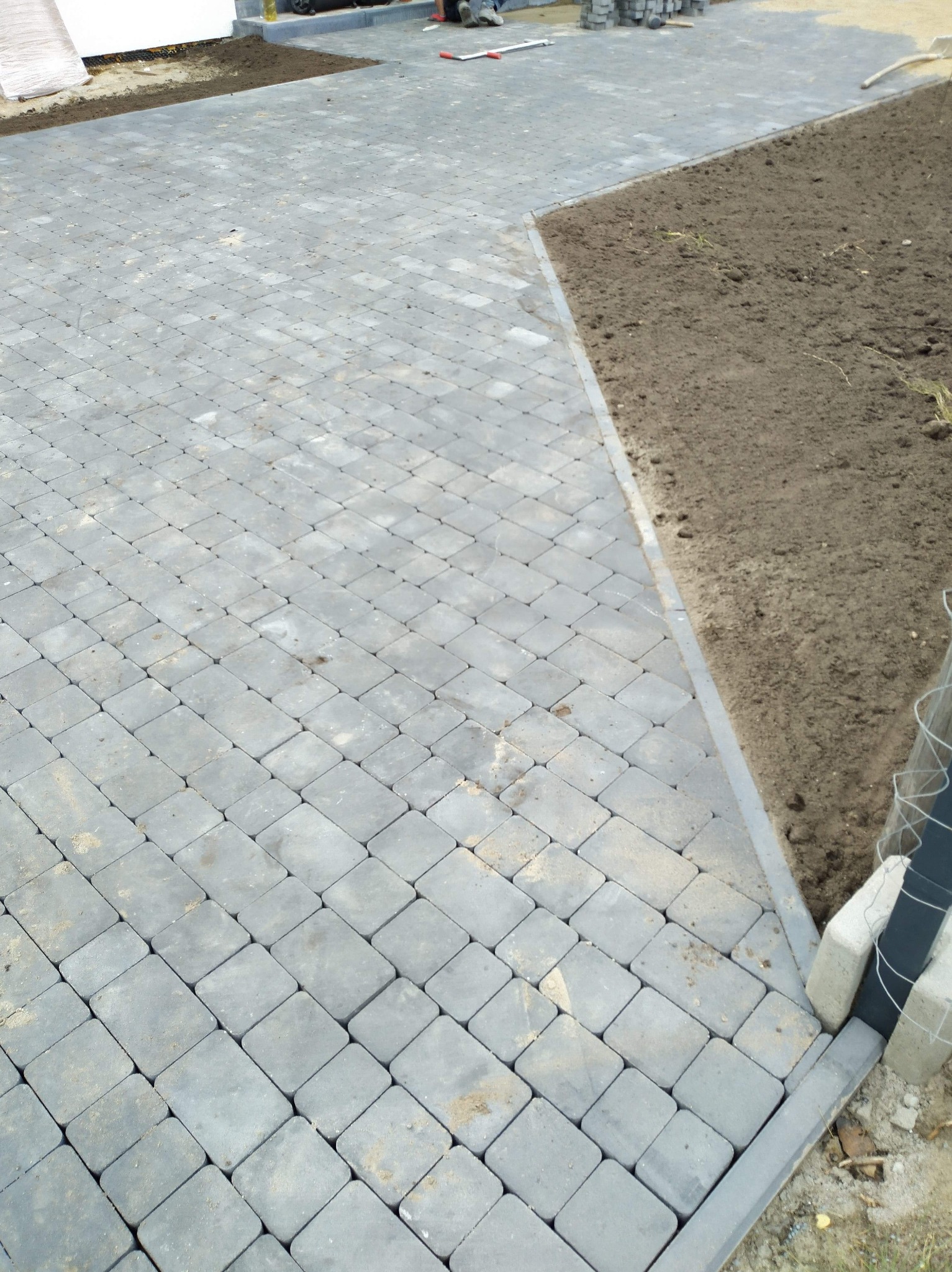 Ułożona szara kostka brukowa w kwadratowy wzór, obok świeżo nawieziona ziemia pod trawnik, widoczne elementy betonowego obrzeża.