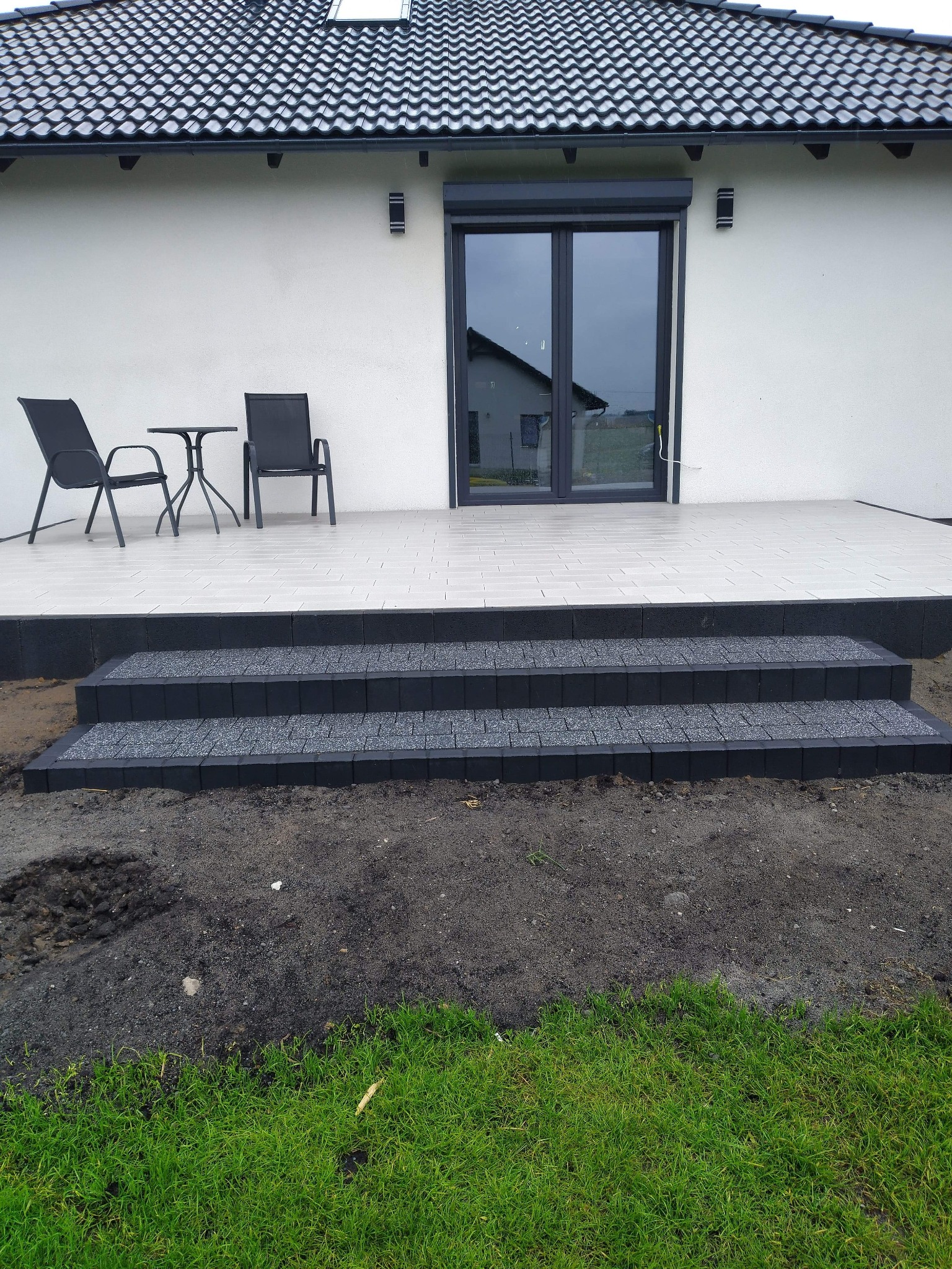 Nowoczesny taras z jasnych płytek z ciemnymi schodami z kostki brukowej, widoczne krzesła i stolik, dom z czarnym dachem w tle.