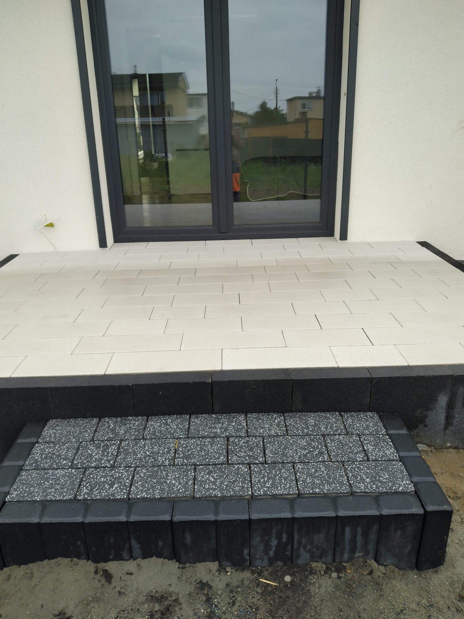 Wykończony taras z jasnych, prostokątnych płyt z ciemnoszarą obwódką i stopniem z szarej kostki brukowej przed dwuskrzydłowymi drzwiami balkonowymi w nowoczesnym domu.