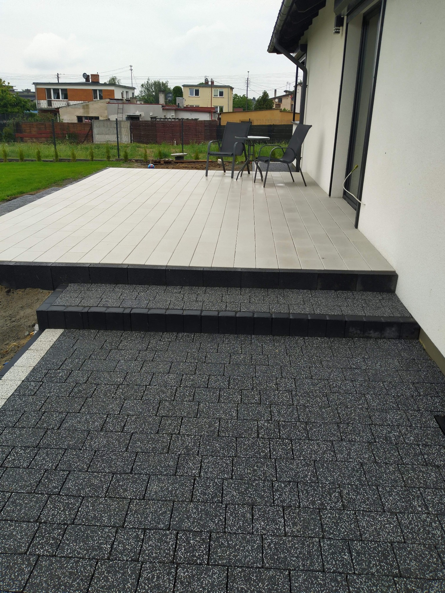 Ułożona ciemnoszara kostka brukowa prowadząca do jasnoszarego tarasu z meblami ogrodowymi przy białym budynku.
