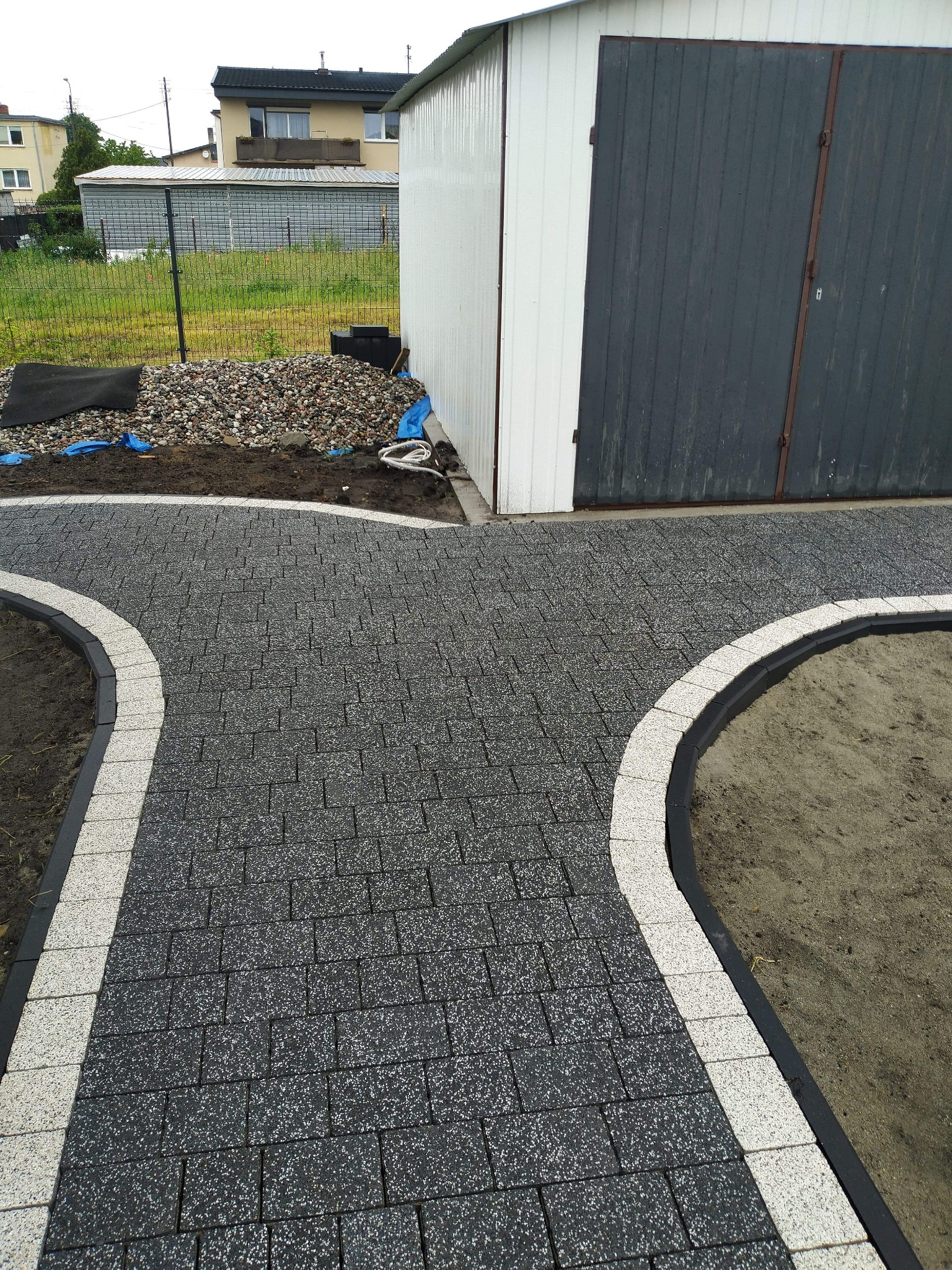 Świeżo ułożona, grafitowa kostka brukowa z jasnymi obrzeżami, rozwidlająca się na dwie ścieżki przed szarym garażem blaszanym i stertą kamieni.