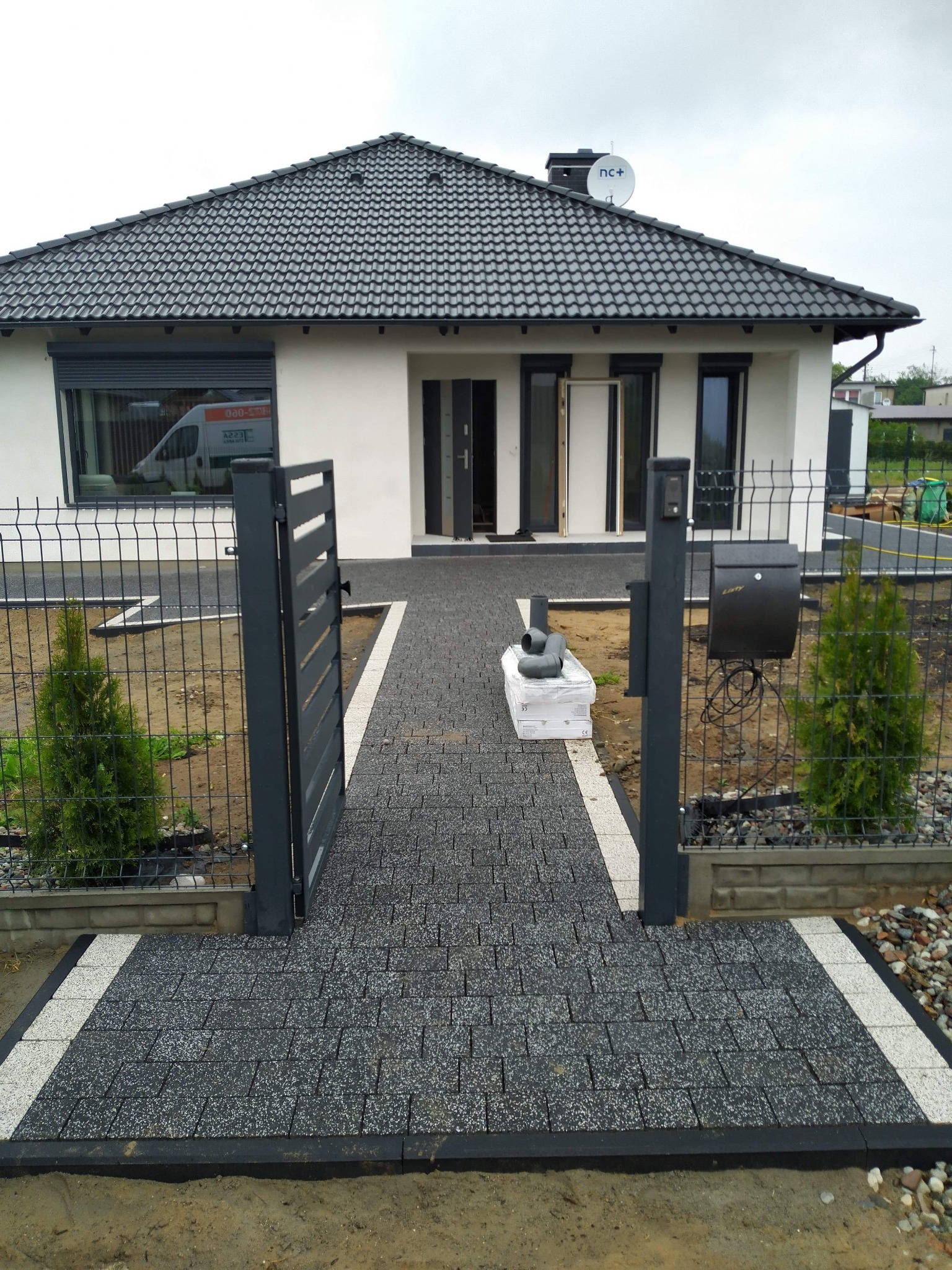 Nowoczesny dom jednorodzinny z ciemnoszarą kostką brukową prowadzącą od furtki do wejścia, obrzeża wykończone jasnymi płytami, widoczny fragment ogrodzenia panelowego.
