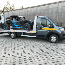 Szary bus laweta z żółtymi pasami transportuje czarno-niebieskiego quada na tle białej naczepy ciężarówki i drzew bez liści.