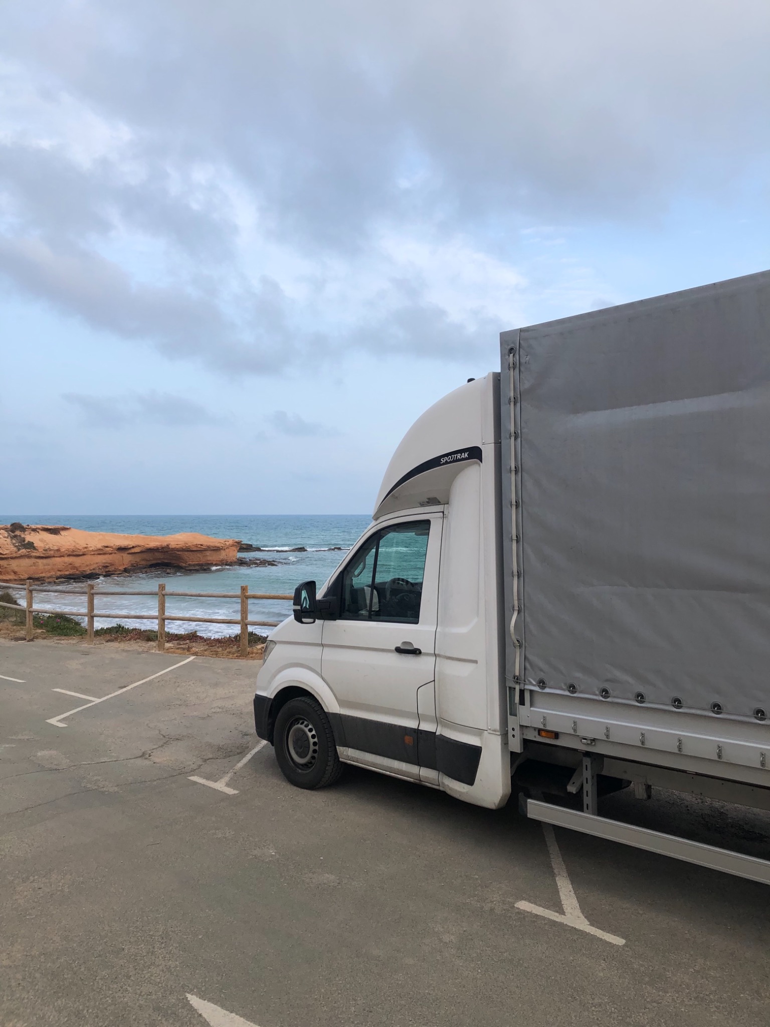 Biały samochód dostawczy typu furgon z plandeką parkuje na parkingu z widokiem na morze i skaliste wybrzeże w pochmurny dzień.
