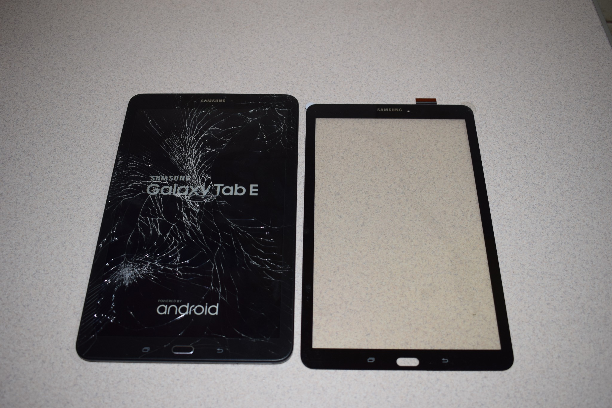 Uszkodzony tablet Samsung Galaxy Tab E z pękniętym ekranem obok nowego, pustego panelu dotykowego na szarym tle.