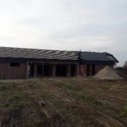 Budowa domu z keramzytu w trakcie: widoczna konstrukcja dachu z częściowo ułożoną dachówką, robotnicy na drabinie przy ciemnym fragmencie dachu, oraz sterta piasku obok budynku.