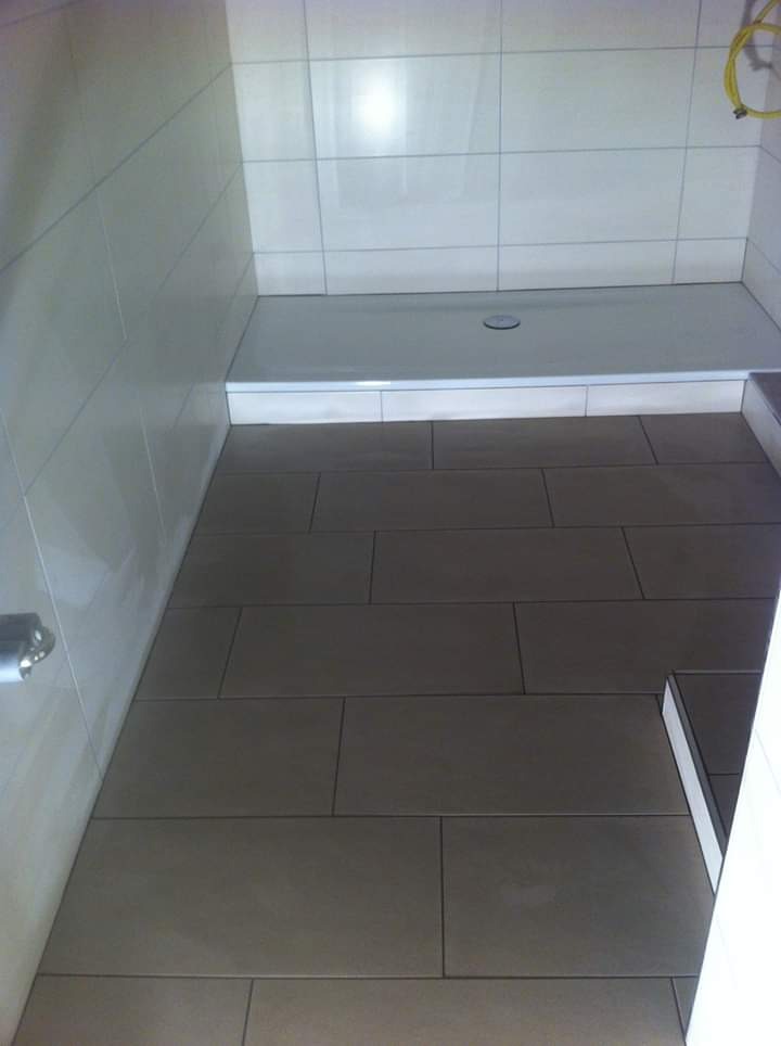 Wyłożony brązowymi płytkami podłogowymi i białymi płytkami ściennymi brodzik z odpływem. Widoczny fragment żółtego kabla.