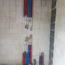 Instalacja wodna w surowym stanie: pionowe rury w izolacji czerwonej i niebieskiej, mocowane do ściany z betonu komórkowego, z widocznymi manometrami i zaworami.