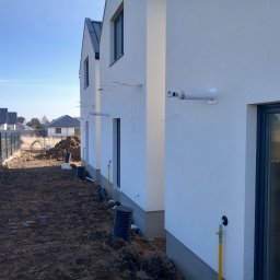 Usługi hydrauliczne - Biały budynek w trakcie instalacji zewnętrznej, widoczne rury i przewody. Ujęcie z ziemi, w tle inne budynki i błękitne niebo. Prace instalacyjne w nowym budownictwie.