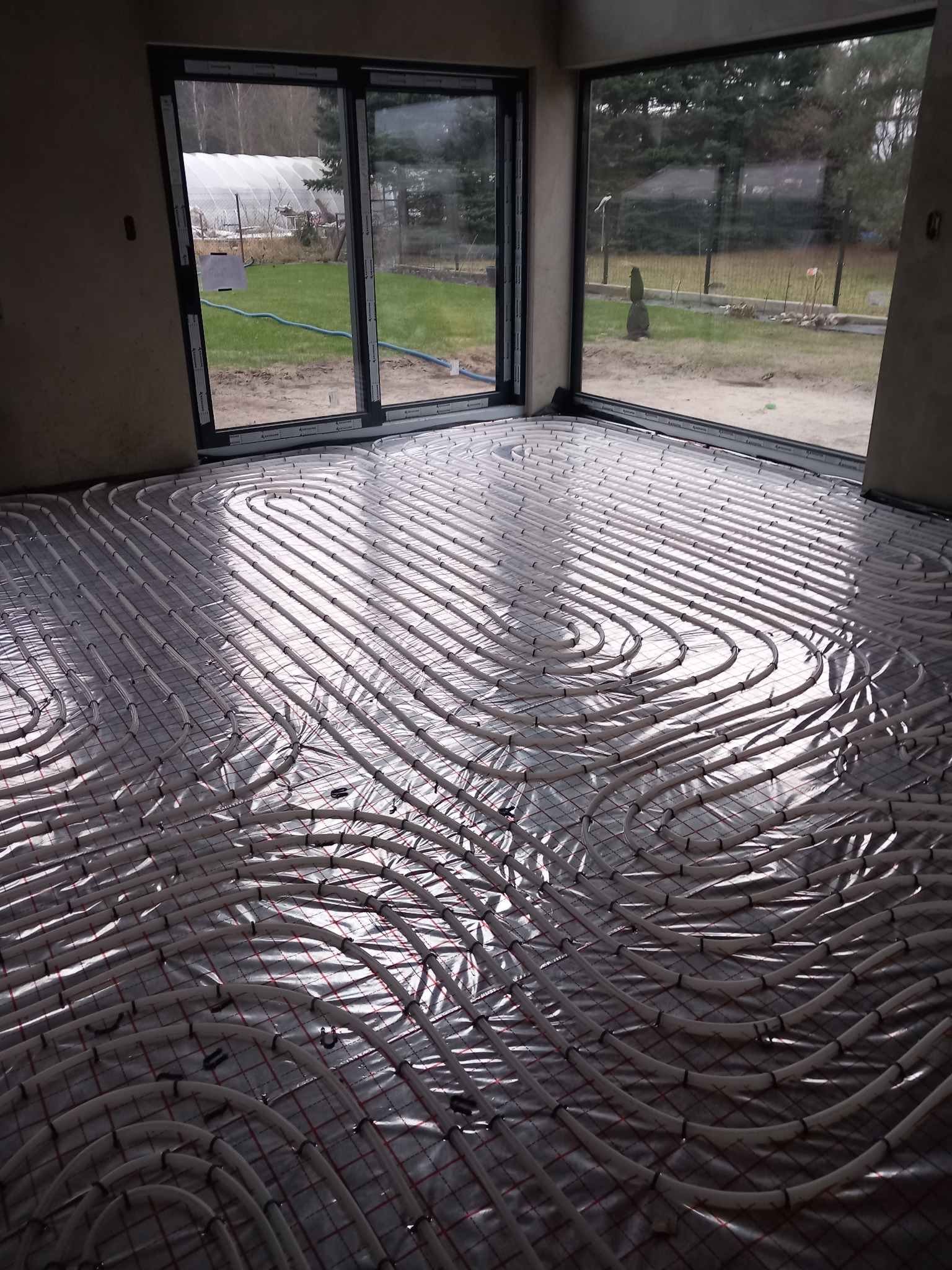 Białe rury ogrzewania podłogowego ułożone na srebrnej folii izolacyjnej w surowym pomieszczeniu z dużymi oknami wychodzącymi na ogród z tunelem foliowym.