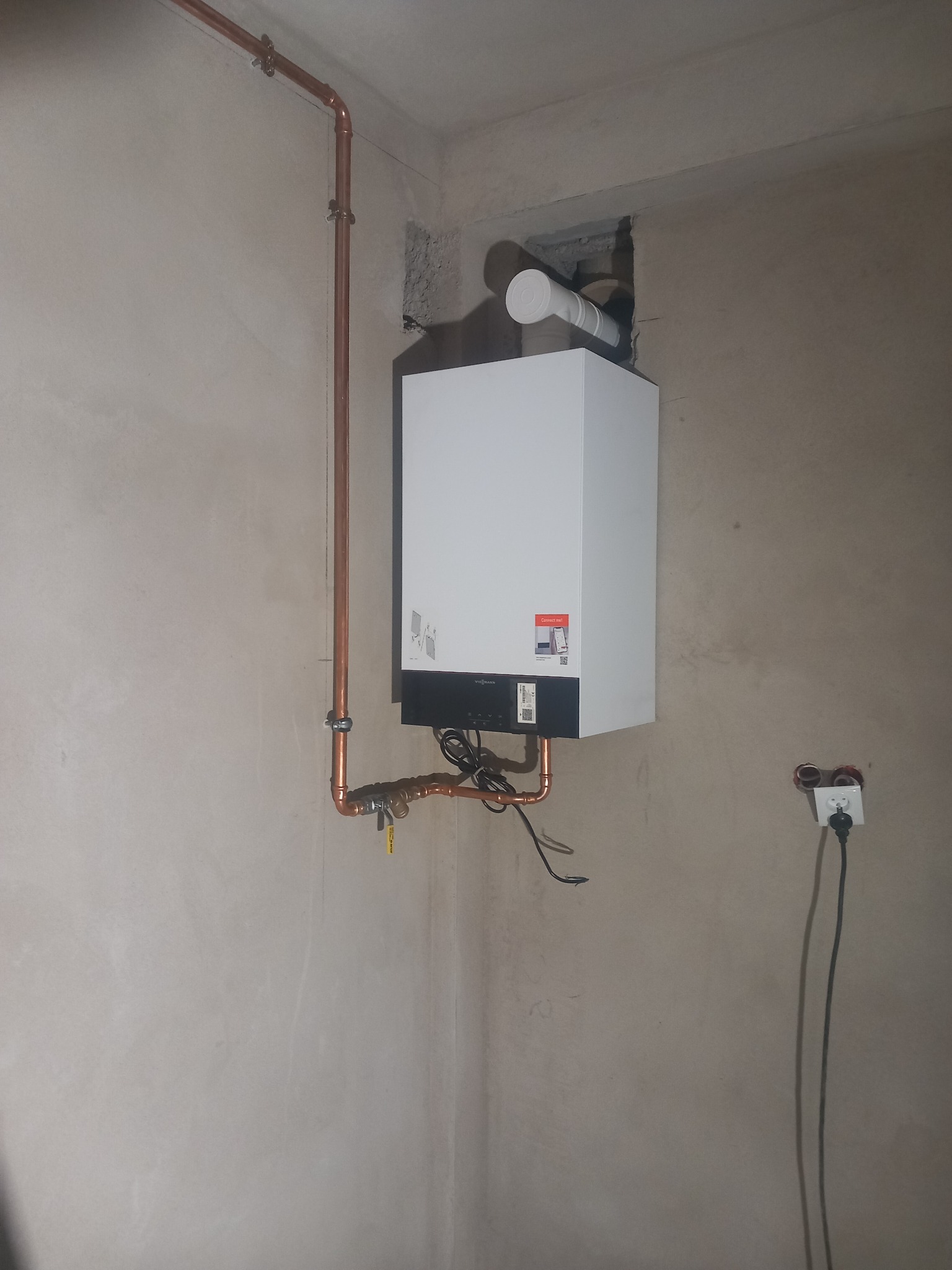 Nowy, biały piec gazowy zamontowany na szarej ścianie z widocznymi miedzianymi rurami, podłączeniem do komina i gniazdkiem elektrycznym. Zwraca uwagę surowy charakter pomieszczenia w trakcie...