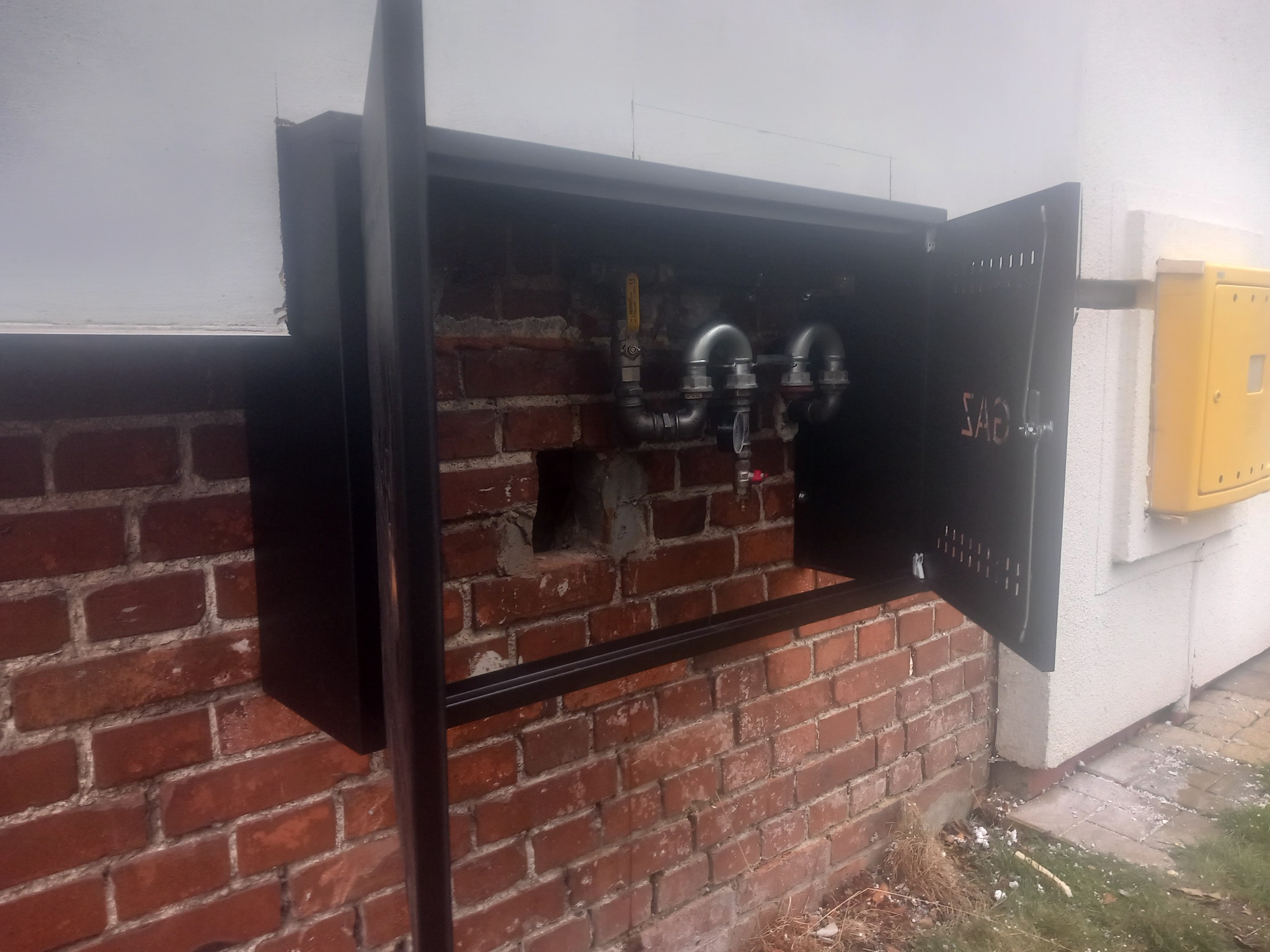 Zewnętrzna skrzynka gazowa z otwartymi drzwiczkami, odsłaniająca metalowe rury i zawory na tle ceglanej ściany. Obok widoczna żółta skrzynka gazowa na białym tynku.