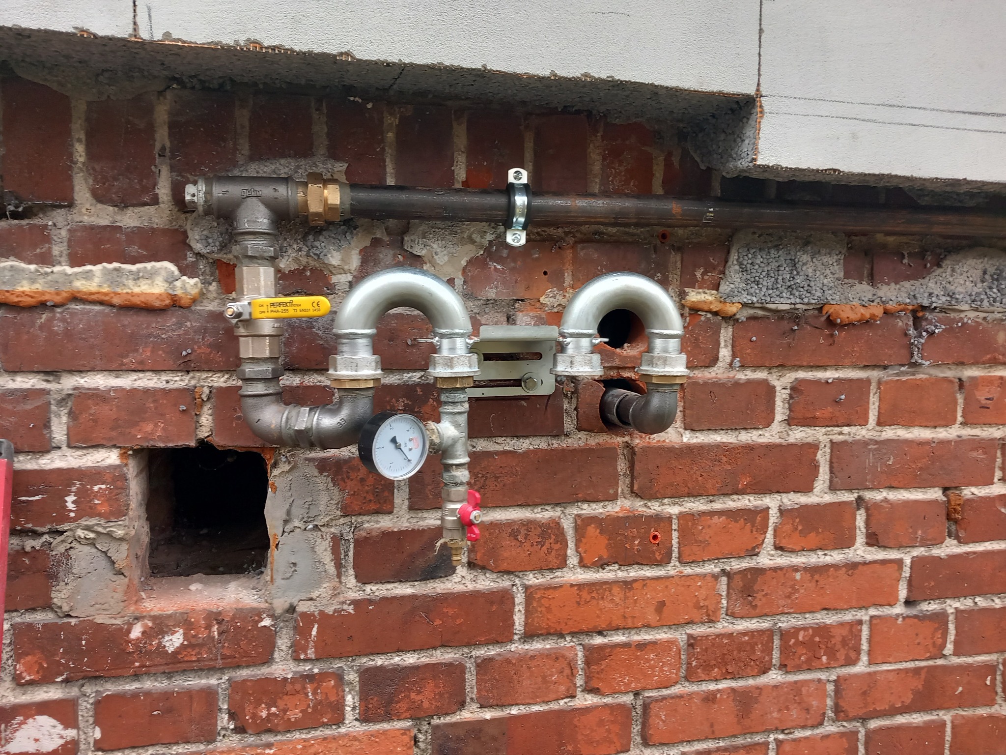 Zewnętrzna instalacja gazowa z rurami, zaworami i manometrem zamontowana na ceglanej ścianie budynku z widocznymi śladami napraw i izolacji.