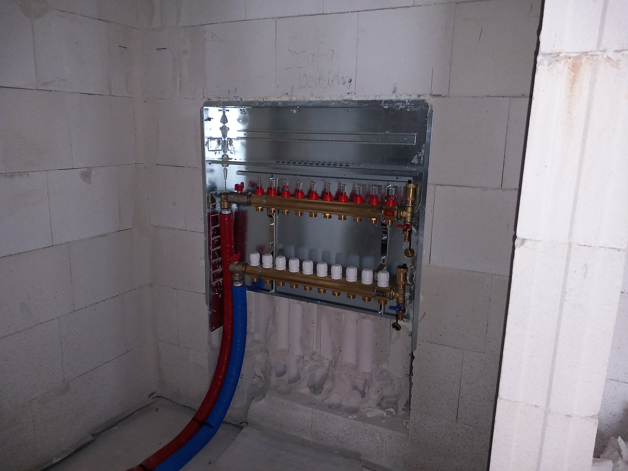 Rozdzielacz ogrzewania podłogowego zamontowany w metalowej szafce, widoczne rury doprowadzające wodę w kolorach czerwonym i niebieskim, ściany z betonu komórkowego z napisem 'Szafa rozdzielaczy'.