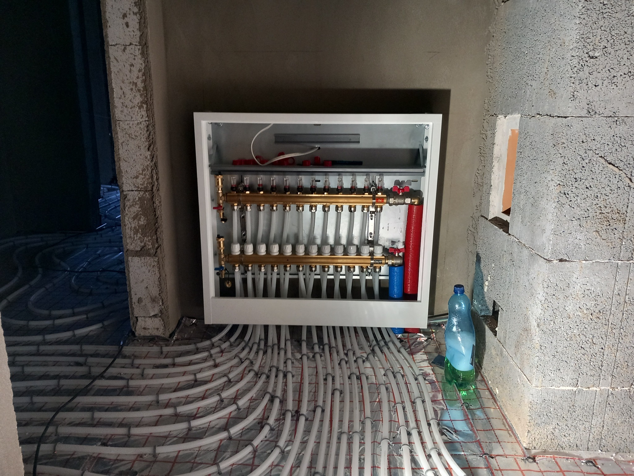 Rozdzielacz ogrzewania podłogowego w białej szafce, widoczne rury rozprowadzone na podłodze, surowy stan wykończenia ścian, butelka z wodą obok ściany.