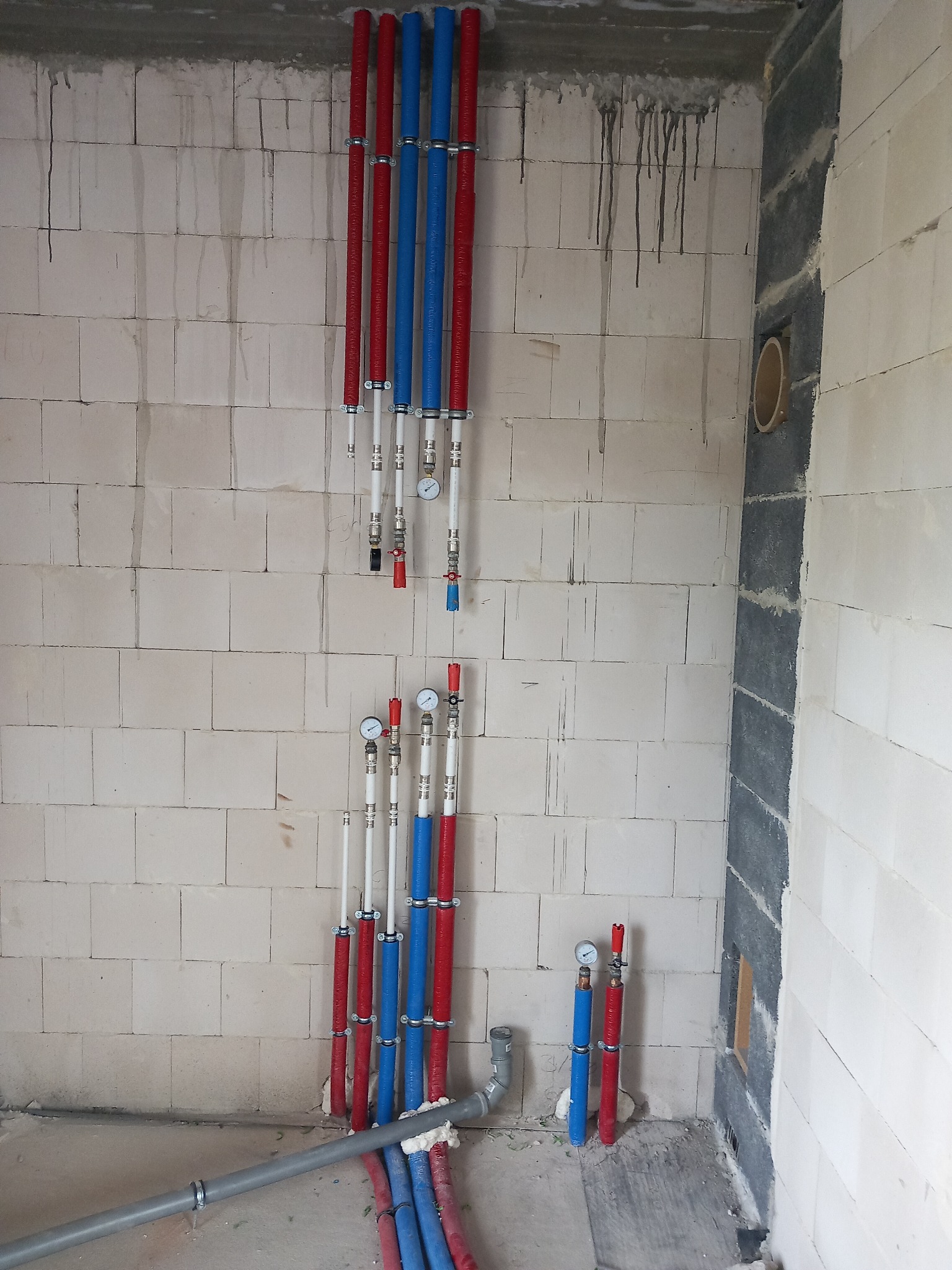 Instalacja wodna w surowym stanie: pionowe rury w izolacji czerwonej i niebieskiej, mocowane do ściany z betonu komórkowego, z widocznymi manometrami i zaworami.