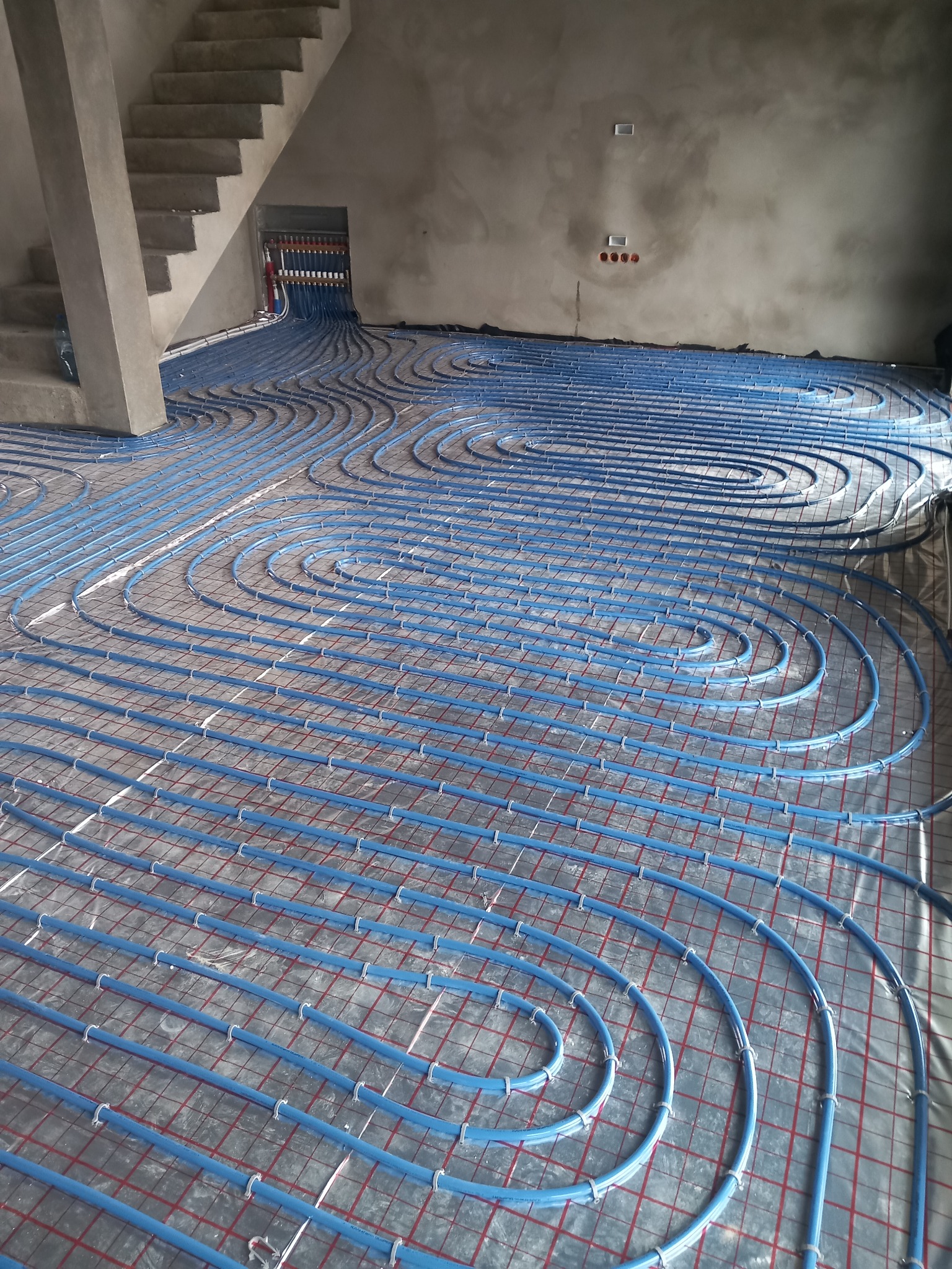Niebieskie rury ogrzewania podłogowego ułożone spiralnie na siatce w surowym wnętrzu z betonowymi schodami i ścianami przed wylewką.
