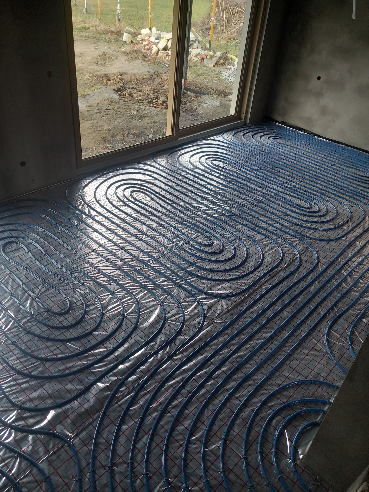 Niebieskie rury ogrzewania podłogowego ułożone na srebrnej folii z czerwoną siatką, widok z góry na surowy stan wykończenia wnętrza z oknem i widokiem na zewnątrz.
