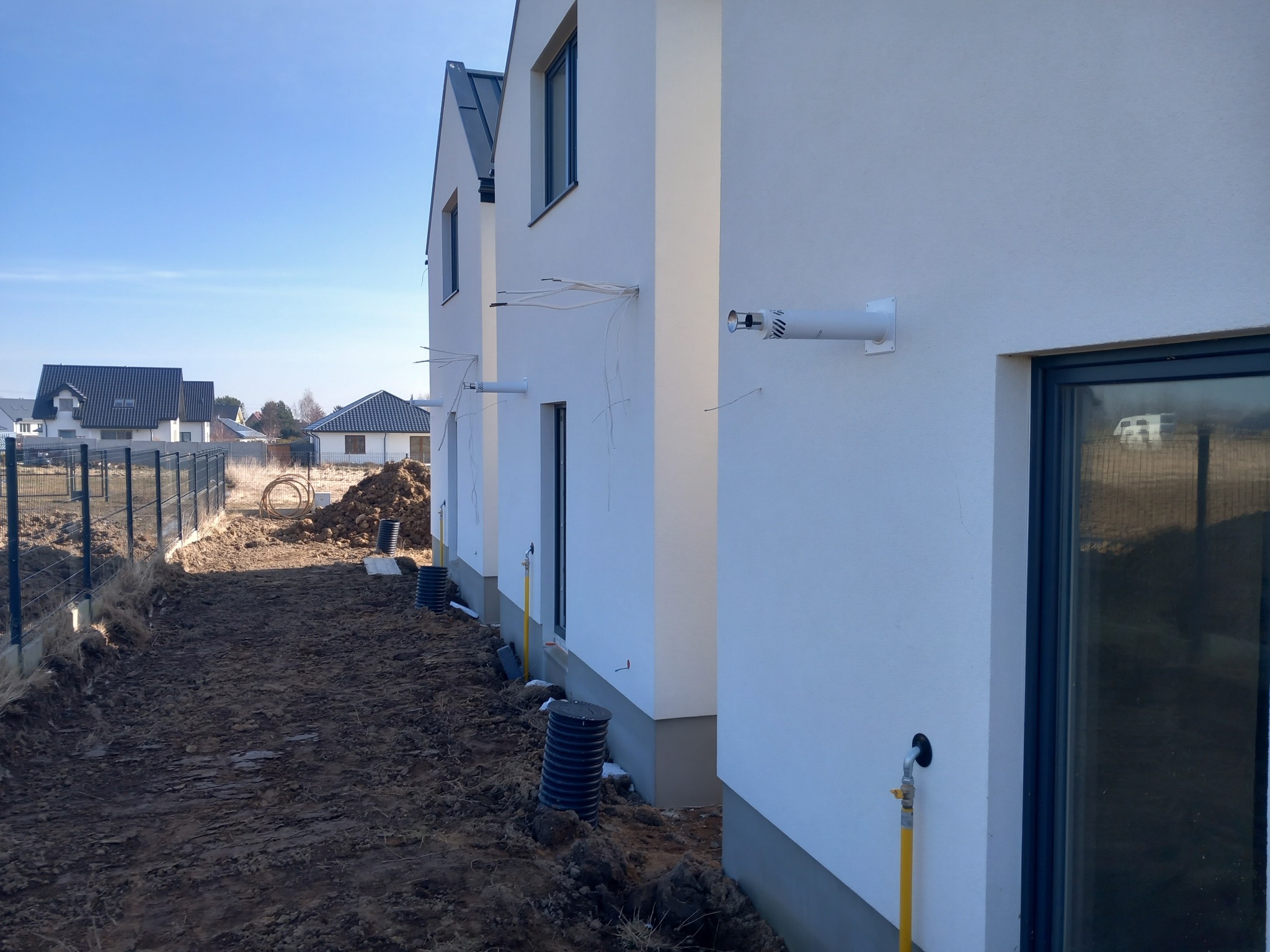 Biały budynek w trakcie instalacji zewnętrznej, widoczne rury i przewody. Ujęcie z ziemi, w tle inne budynki i błękitne niebo. Prace instalacyjne w nowym budownictwie.