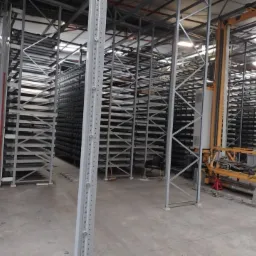 Wnętrze hali magazynowej z wysokimi, metalowymi regałami, widoczne elementy konstrukcji i system automatycznego transportu towarów.