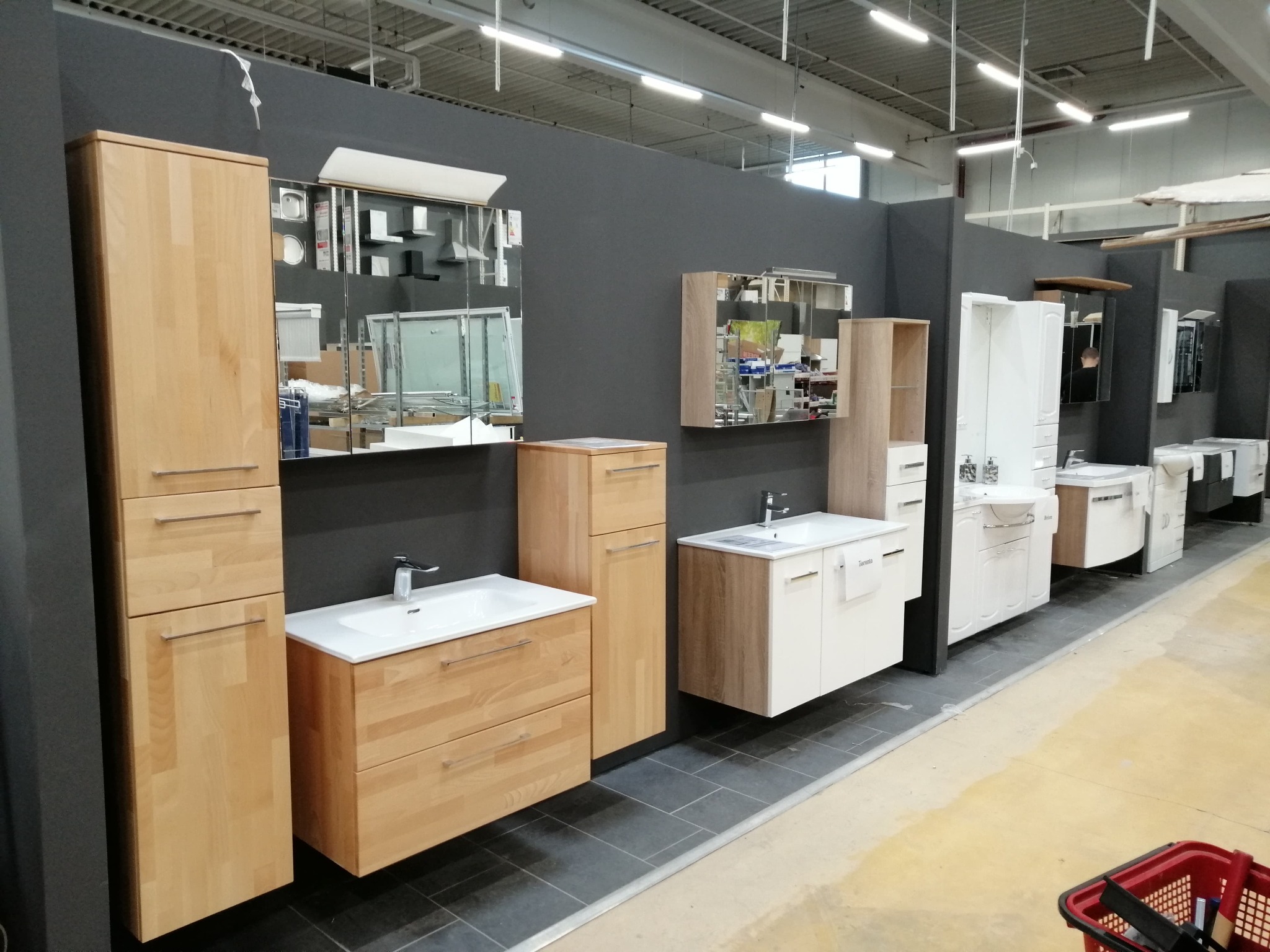 Ekspozycja mebli łazienkowych w sklepie: szafki pod umywalki z lustrami, różne style i wykończenia, oświetlenie sufitowe, widoczny koszyk zakupowy w tle.