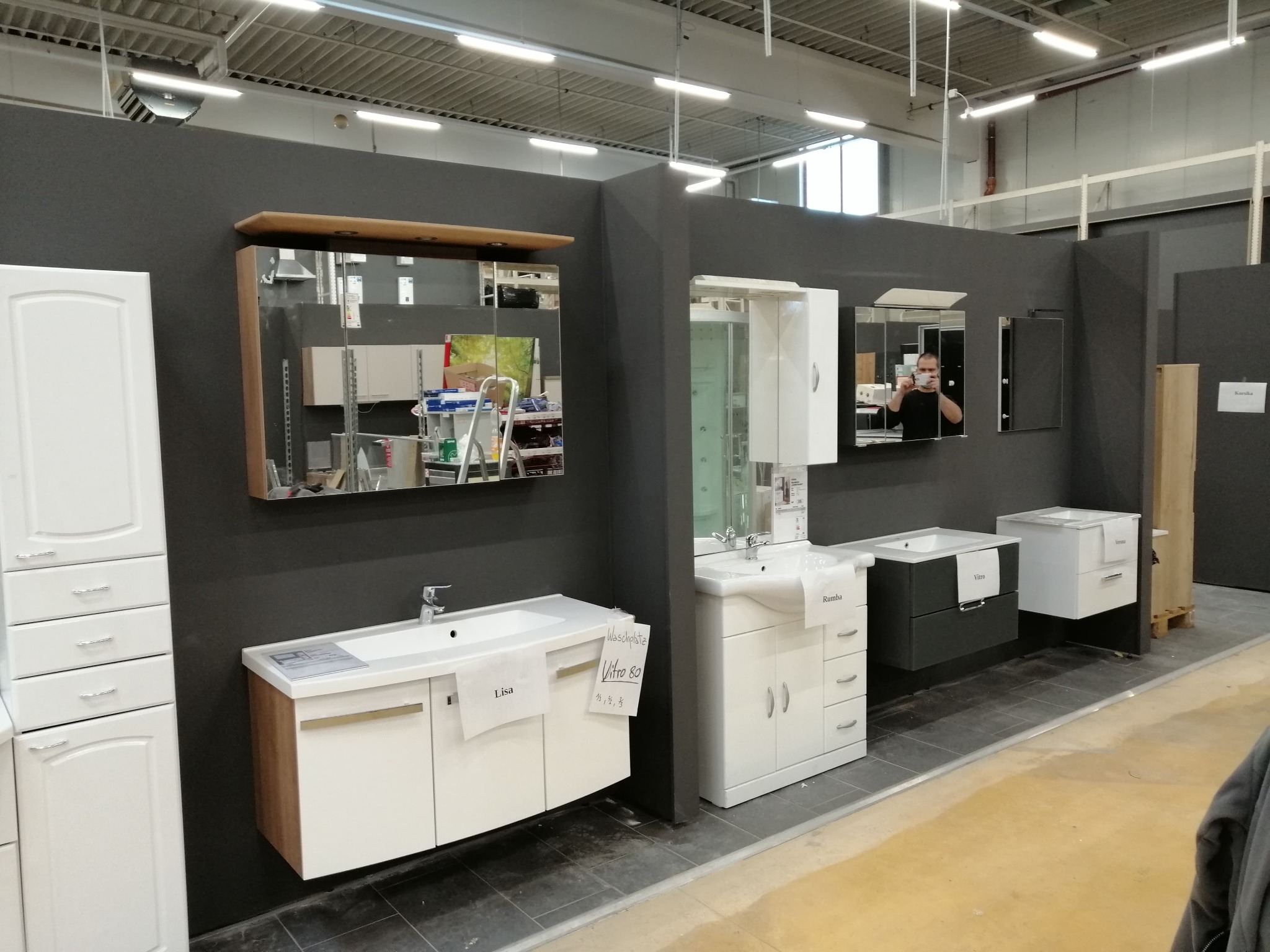 Ekspozycja różnych modeli szafek łazienkowych z umywalkami i lustrami w sklepie, widoczne ceny i nazwy modeli na kartkach.