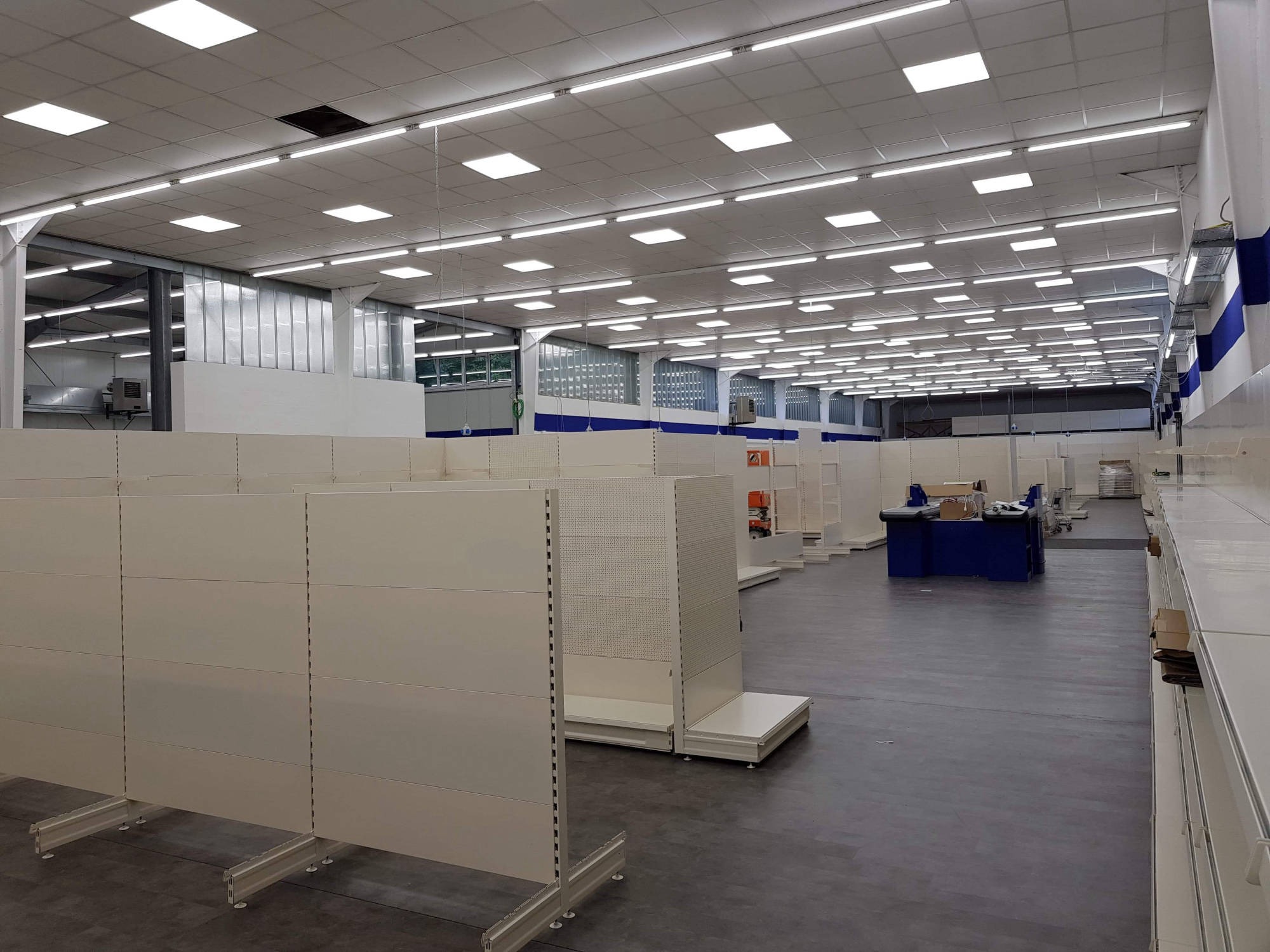 Montaż regałów magazynowych w nowej hali sklepu, widoczne puste białe regały z perforowanymi ścianami, równomierne oświetlenie sufitowe.