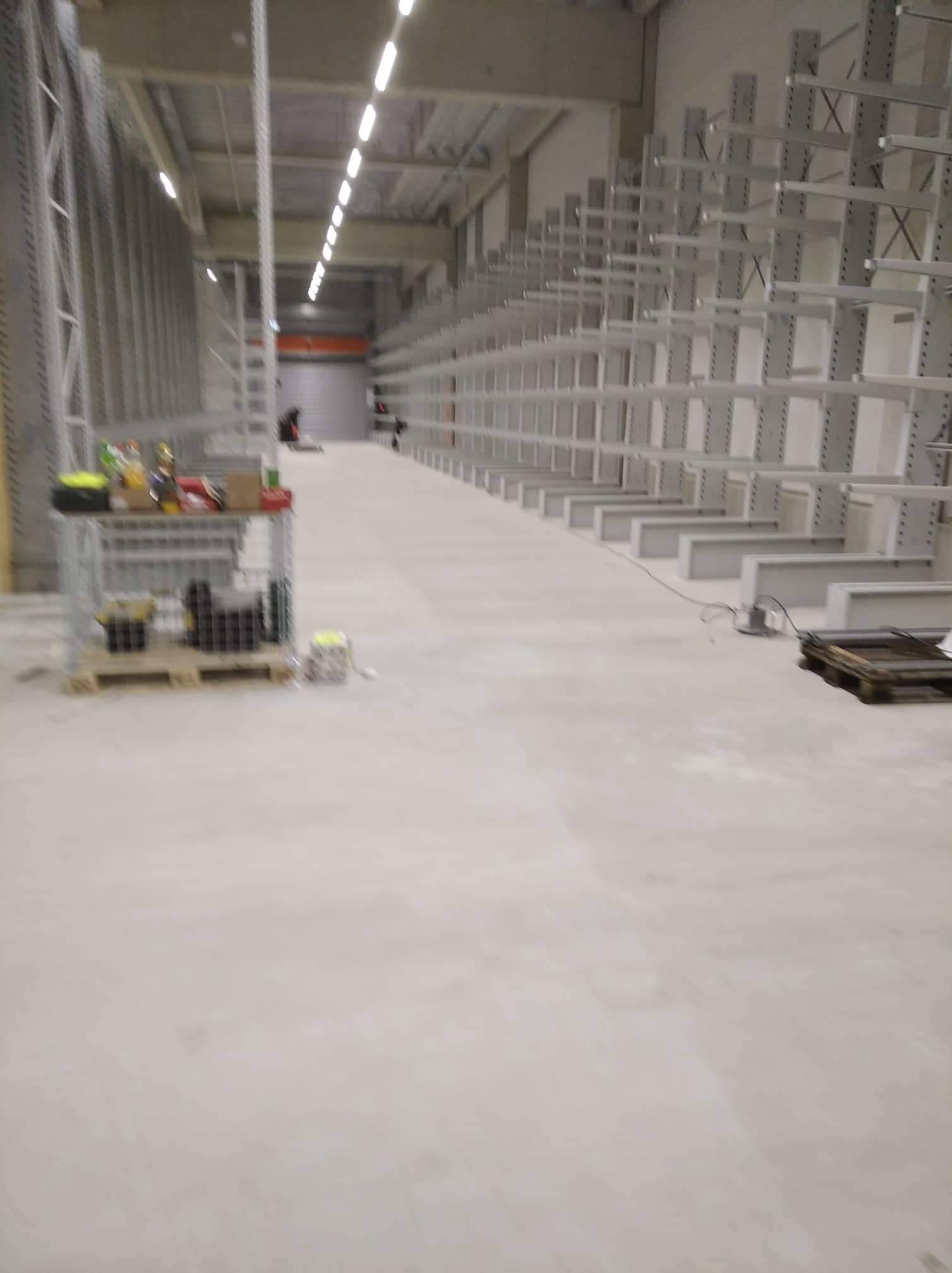 Długi magazyn z rzędami metalowych regałów bez półek, widoczne narzędzia i materiały na palecie. W tle pracownik magazynu.