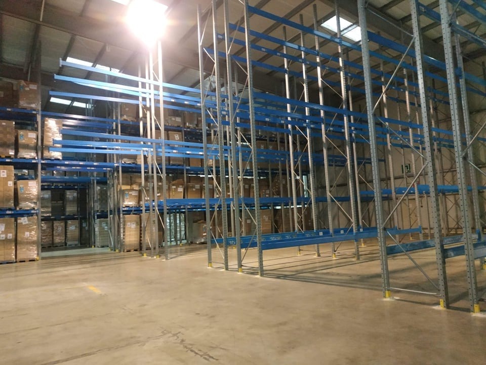 Wnętrze magazynu z częściowo zapełnionymi regałami paletowymi w kolorze niebieskim i srebrnym, oświetlone światłem słonecznym wpadającym przez okna dachowe.