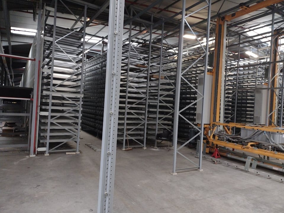 Wnętrze hali magazynowej z wysokimi, metalowymi regałami, widoczne elementy konstrukcji i system automatycznego transportu towarów.