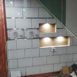 Wykończenie łazienki na poddaszu: białe płytki z motywem kwiatowym, wnęki z oświetleniem, widoczna konstrukcja z płyty kartonowo-gipsowej i narzędzie do cięcia płytek.