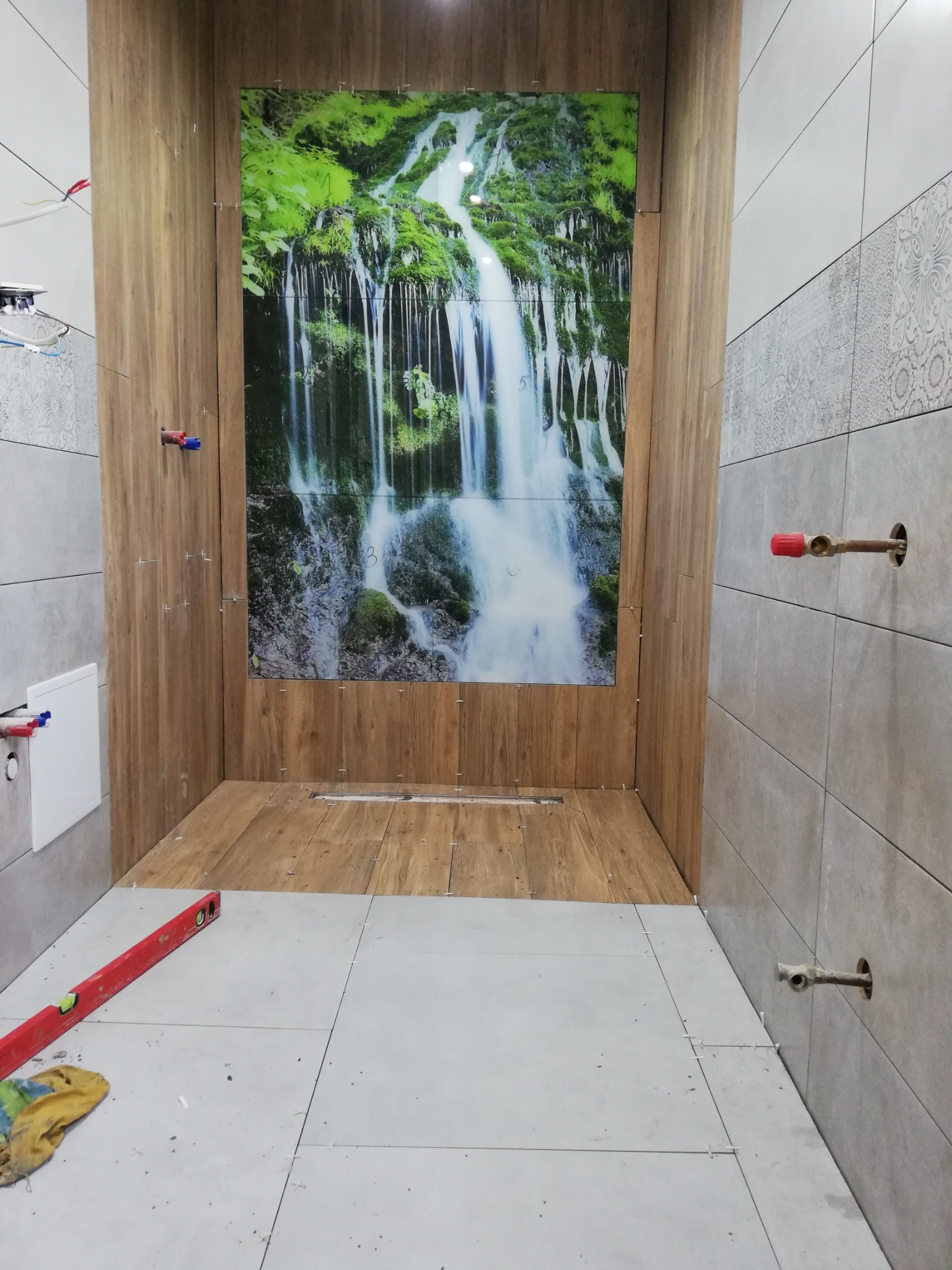 Wnętrze łazienki w trakcie wykańczania: ułożone płytki podłogowe i ścienne, imitacja drewna na ścianie prysznica z grafiką wodospadu, widoczna poziomica i wystające rury.