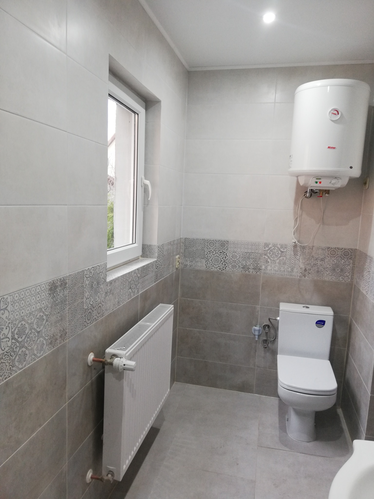 Wykończona łazienka z szarymi płytkami, dekoracyjnym pasem z wzorami, białym grzejnikiem, podgrzewaczem wody i toaletą.