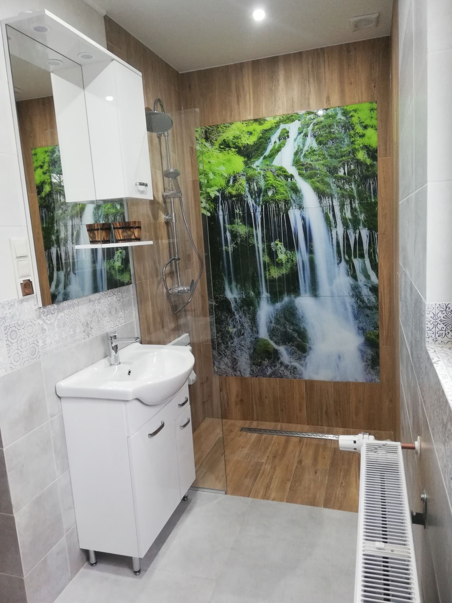 Nowoczesna łazienka z białą szafką pod umywalką, kabiną prysznicową typu walk-in i panelem dekoracyjnym z motywem wodospadu.