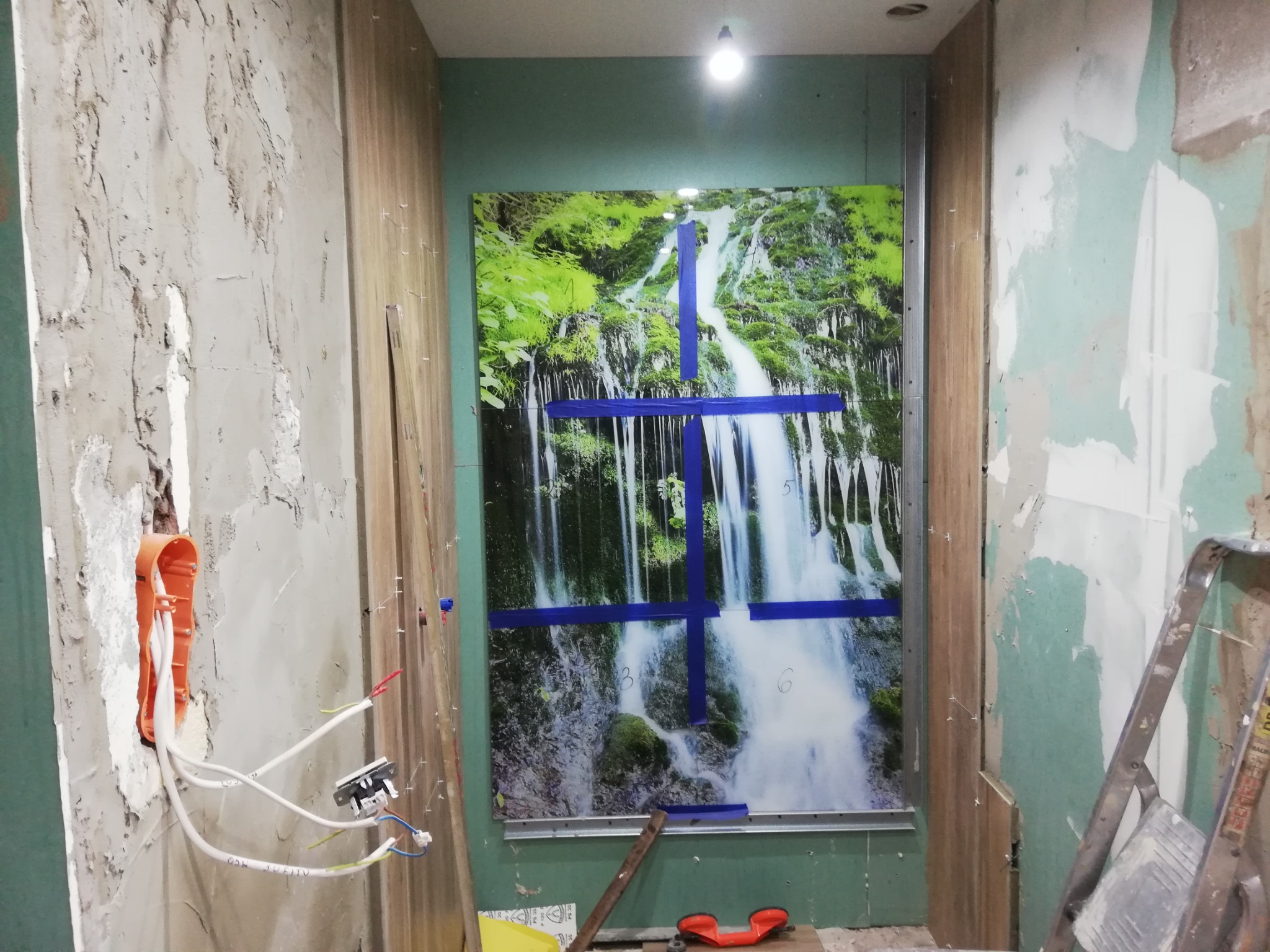 Montaż panelu dekoracyjnego z motywem wodospadu, przytwierdzonego niebieską taśmą, widoczne przewody elektryczne i fragment ściany w trakcie prac wykończeniowych.
