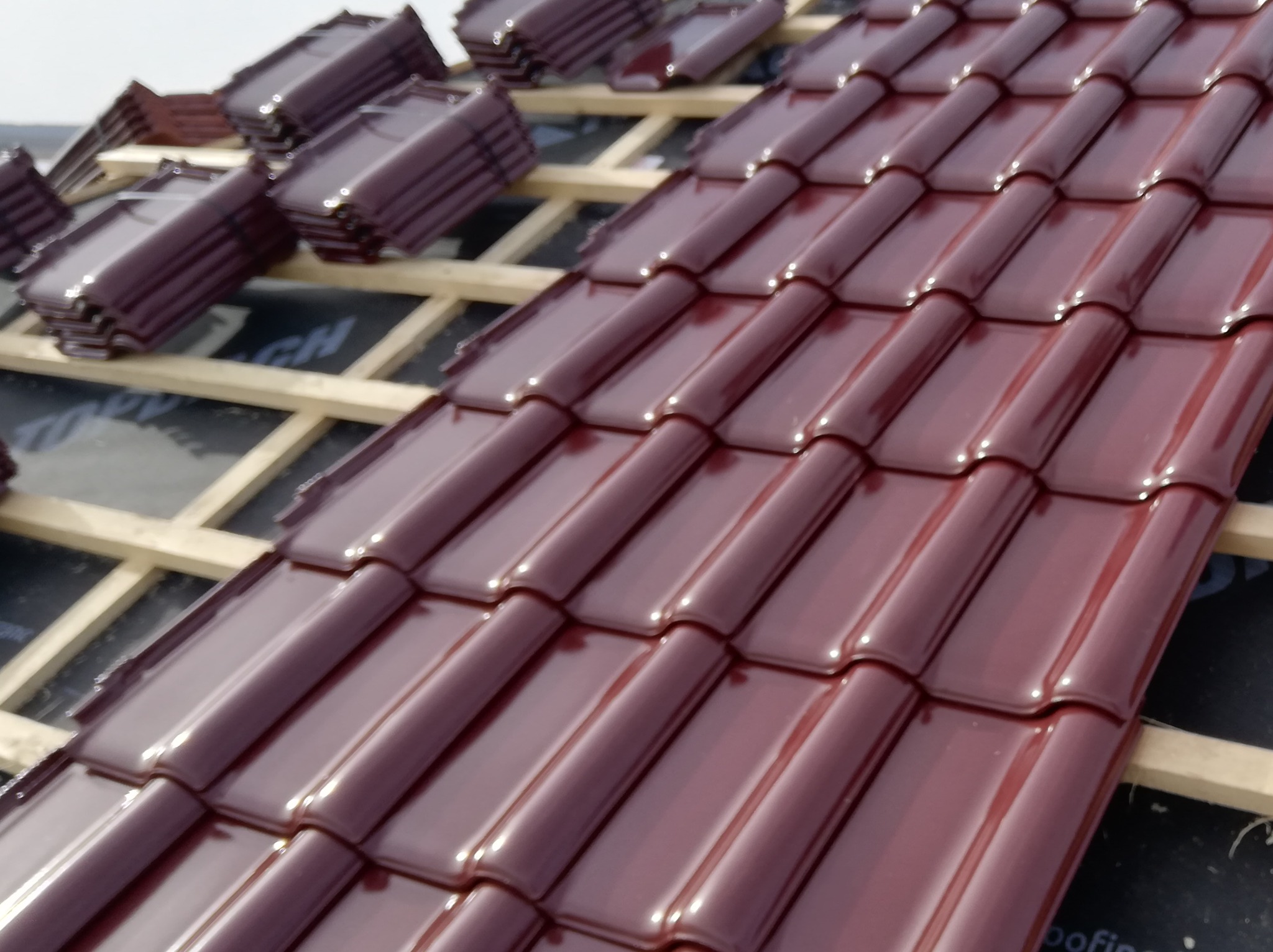 Układanie bordowej, błyszczącej dachówki ceramicznej na łatach drewnianych, widoczne membrany dachowe.