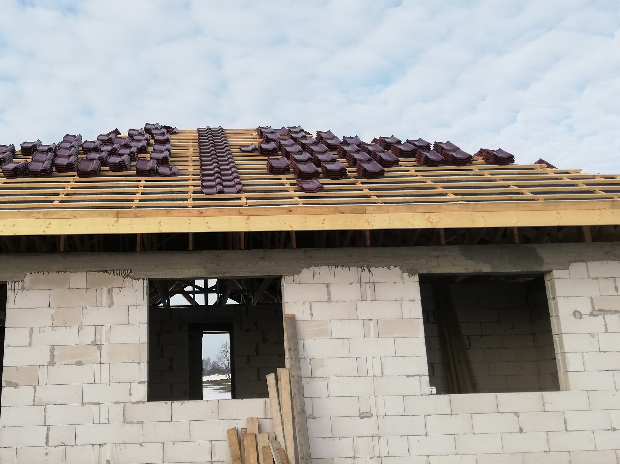 Budowa domu jednorodzinnego w trakcie: białe bloczki, drewniana konstrukcja dachu z ułożoną częścią brązowej dachówki ceramicznej, widok na zimowy krajobraz przez otwory okienne.