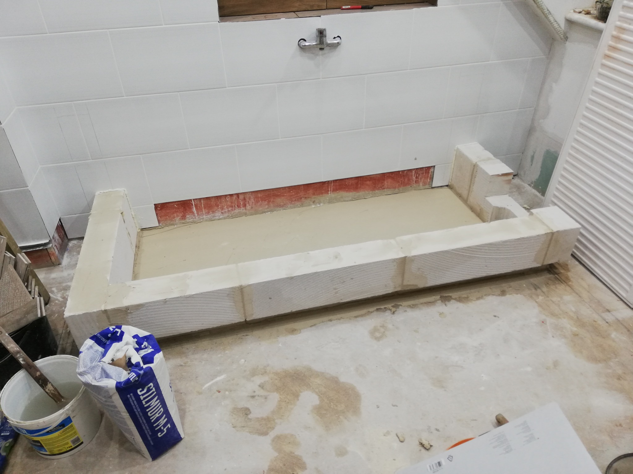 Przygotowanie pod zabudowę wanny z bloczków, z widocznym workiem zaprawy Silmur M-5 i białymi płytkami na ścianie w tle.
