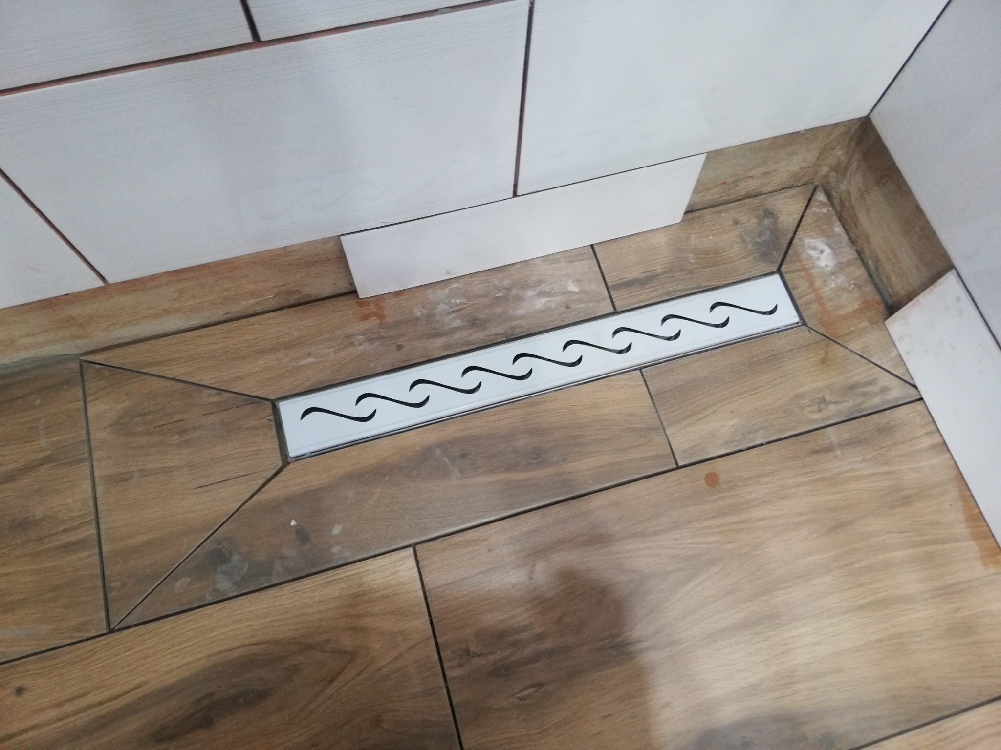 Ułożona podłoga z płytek imitujących drewno z odpływem liniowym w kabinie prysznicowej, białe płytki ścienne w tle.